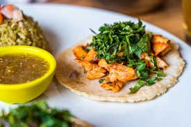 A dish featuring a taco on a white plate, topped with grilled chicken pieces and fresh green cilantro. A yellow bowl containing green salsa sits next to the taco. There is also a scoop of green rice with vegetables in the background.