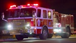 A fire truck is seen at night with its lights flashing. The vehicle has a large grille at the front and multiple compartments along the sides with hoses and equipment. The bright lights on top cast a red and white glow.