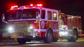 A fire truck is seen at night with its lights flashing. The vehicle has a large grille at the front and multiple compartments along the sides with hoses and equipment. The bright lights on top cast a red and white glow.
