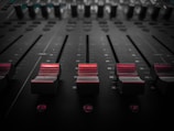 A close-up of a sound mixing console, featuring several dark sliders with red accents across their surfaces. The sliders are aligned horizontally on a black panel, with numbered markings indicating positions. The lighting casts shadows, creating a moody atmosphere.