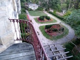 A beautifully landscaped garden with a stone walkway meandering through well-manicured lawns and plant beds. The garden features colorful flower arrangements and a white gazebo in the distance, surrounded by lush greenery and mature trees. A prominent red railing is visible in the foreground, likely part of a balcony overlooking the garden.