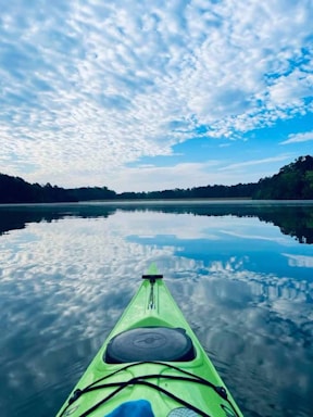 A lime green kayak floats on a calm lake under a sky filled with soft, scattered clouds. The water reflects the sky, creating a mirror-like surface. Dense, dark green trees line the horizon, contrasting with the bright blue and white of the sky.