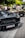 A row of luxury sports cars is parked on a street, with people gathered around them. The cars have sleek, aerodynamic designs, and the one in the foreground has a customized license plate. The scene is set against a backdrop of trees and a bright, sunny atmosphere.