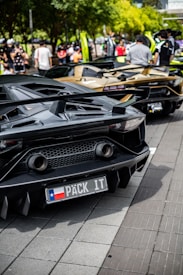 A row of luxury sports cars is parked on a street, with people gathered around them. The cars have sleek, aerodynamic designs, and the one in the foreground has a customized license plate. The scene is set against a backdrop of trees and a bright, sunny atmosphere.