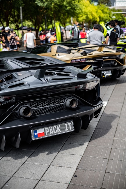 A row of luxury sports cars is parked on a street, with people gathered around them. The cars have sleek, aerodynamic designs, and the one in the foreground has a customized license plate. The scene is set against a backdrop of trees and a bright, sunny atmosphere.