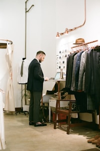 A man is standing in a well-lit interior space, working on a laptop placed on a desk. The surroundings include a neatly organized clothing rack with suits, hats, and shoes. The decor includes metallic accents and a simplistic, modern aesthetic.
