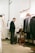 A man is standing in a well-lit interior space, working on a laptop placed on a desk. The surroundings include a neatly organized clothing rack with suits, hats, and shoes. The decor includes metallic accents and a simplistic, modern aesthetic.