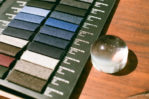 A collection of fabric swatches arranged in rows, displaying a variety of colors and textures, is positioned on a wooden surface. Each swatch has text printed beside it, possibly detailing fabric information. Nearby, a clear glass paperweight with a slight distortion from the light is placed.