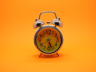 A classic-style alarm clock with a shiny metal frame and two bells on top stands against a bright orange background. The clock face features bold black numbers, with colorful red and blue numbers highlighting the three and nine positions. The hands of the clock are black and point at 10:09.