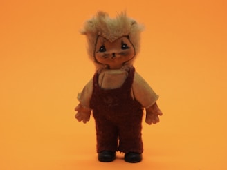 A small stuffed toy designed to look like an animal, possibly a cat, wearing a tan colored shirt and dark maroon overalls. The toy has a fuzzy tail and its face has whiskers, large eyes, and a simple nose and mouth. The background is a solid bright orange.
