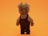 A small stuffed toy designed to look like an animal, possibly a cat, wearing a tan colored shirt and dark maroon overalls. The toy has a fuzzy tail and its face has whiskers, large eyes, and a simple nose and mouth. The background is a solid bright orange.