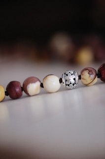 A close-up of a string of beads resting on a flat surface, featuring various earthy tones and textures. One of the beads is metallic with an intricate design, contrasting with the matte finish of the surrounding beads.