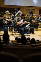 A symphony orchestra performs on stage with musicians playing various string and brass instruments. The musicians are dressed in formal black attire, focusing intently on their music. The setting is an elegant concert hall with rows of wooden seats visible in the foreground, occupied by an audience.