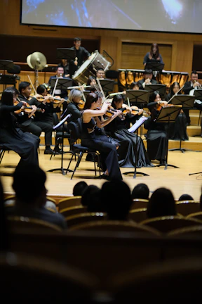 A symphony orchestra performs on stage with musicians playing various string and brass instruments. The musicians are dressed in formal black attire, focusing intently on their music. The setting is an elegant concert hall with rows of wooden seats visible in the foreground, occupied by an audience.
