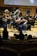 A symphony orchestra performs on stage with musicians playing various string and brass instruments. The musicians are dressed in formal black attire, focusing intently on their music. The setting is an elegant concert hall with rows of wooden seats visible in the foreground, occupied by an audience.