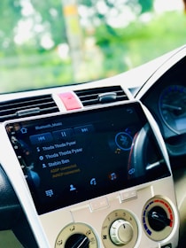 A car's dashboard display screen shows a music player interface with the song 'Thoda Thoda Pyaar' by Stebin Ben. The dashboard features various controls for air conditioning and audio settings. The screen indicates Bluetooth connectivity.