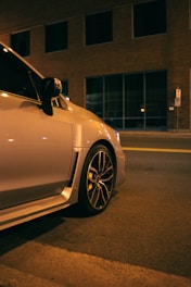 A close-up view of the back half of a modern car parked on a street at night. The car has black alloy wheels with yellow brake calipers. The street is dimly lit, and the backdrop includes a large building with glass windows. The overall lighting suggests a warm, yellowish glow.