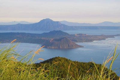 A scenic view of a volcanic island surrounded by a serene body of water, with lush greenery and tall grasses in the foreground. The background features a larger mountainous landscape under a clear sky.