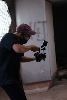 A person wearing a cap and glasses is holding a camera and seems to be in the midst of filming or taking photos in a dimly lit indoor space. The background features a large window with light filtering through and a concrete wall with some graffiti.