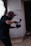 A person wearing a cap and glasses is holding a camera and seems to be in the midst of filming or taking photos in a dimly lit indoor space. The background features a large window with light filtering through and a concrete wall with some graffiti.