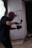 A person wearing a cap and glasses is holding a camera and seems to be in the midst of filming or taking photos in a dimly lit indoor space. The background features a large window with light filtering through and a concrete wall with some graffiti.