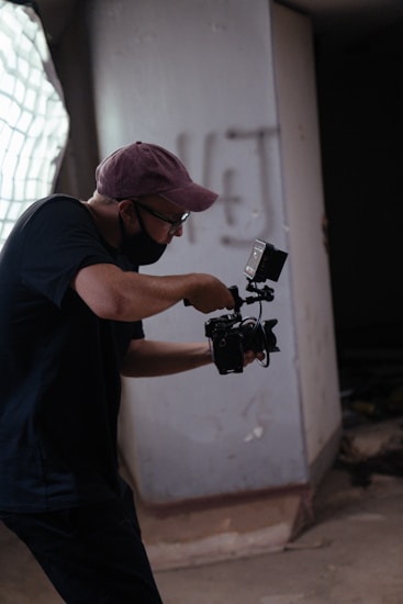 A person wearing a cap and glasses is holding a camera and seems to be in the midst of filming or taking photos in a dimly lit indoor space. The background features a large window with light filtering through and a concrete wall with some graffiti.
