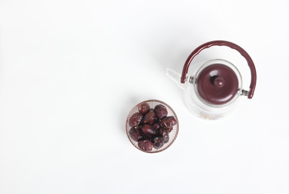 A clear glass teapot with a maroon handle and lid is placed adjacent to a small bowl of dried dates on a white surface.