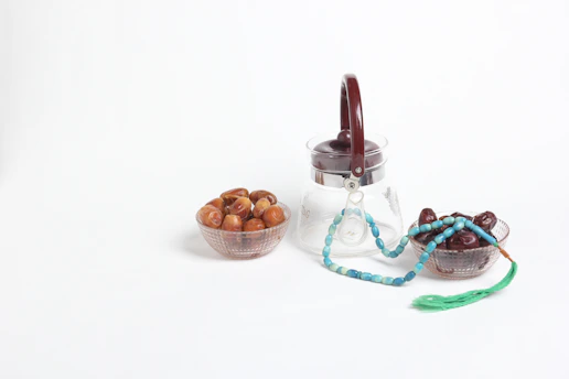 A clear glass jar with a dark brown lid is placed alongside two small bowls filled with dates. A turquoise prayer bead string with a green tassel lies draped over the edge of the right bowl.