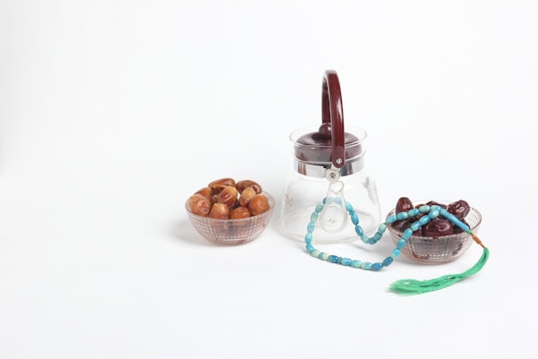 A clear glass jar with a dark brown lid is placed alongside two small bowls filled with dates. A turquoise prayer bead string with a green tassel lies draped over the edge of the right bowl.