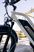A close-up view of a white electric bicycle with a prominent battery pack and branding on the frame. The setting includes a bridge in the background and clear blue skies, creating a bright and inviting atmosphere.