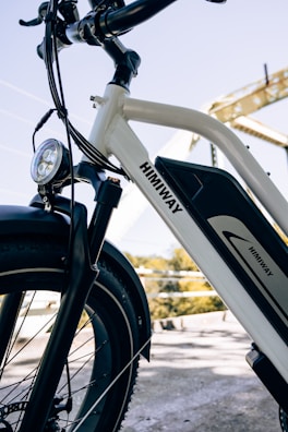 A close-up view of a white electric bicycle with a prominent battery pack and branding on the frame. The setting includes a bridge in the background and clear blue skies, creating a bright and inviting atmosphere.