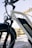 A close-up view of a white electric bicycle with a prominent battery pack and branding on the frame. The setting includes a bridge in the background and clear blue skies, creating a bright and inviting atmosphere.