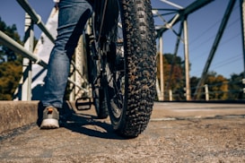 A close-up view of a large mountain bike tire on a concrete bridge. The rider is wearing blue jeans and casual shoes. The background consists of metal bridge railings and distant trees with autumn foliage.