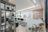 A modern and stylish beauty salon interior featuring wooden accents and a sleek design. The salon has a spacious layout with multiple salon chairs and mirrors under warm pendant lighting. Plants are used as decorative elements, adding a fresh and inviting atmosphere. The reception area has a wooden backdrop with the salon's name displayed, and there's a mix of contemporary furniture and decor.