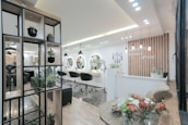 A modern and stylish beauty salon interior featuring wooden accents and a sleek design. The salon has a spacious layout with multiple salon chairs and mirrors under warm pendant lighting. Plants are used as decorative elements, adding a fresh and inviting atmosphere. The reception area has a wooden backdrop with the salon's name displayed, and there's a mix of contemporary furniture and decor.