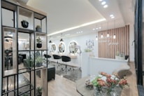 A modern and stylish beauty salon interior featuring wooden accents and a sleek design. The salon has a spacious layout with multiple salon chairs and mirrors under warm pendant lighting. Plants are used as decorative elements, adding a fresh and inviting atmosphere. The reception area has a wooden backdrop with the salon's name displayed, and there's a mix of contemporary furniture and decor.