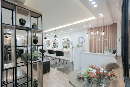A modern and stylish beauty salon interior featuring wooden accents and a sleek design. The salon has a spacious layout with multiple salon chairs and mirrors under warm pendant lighting. Plants are used as decorative elements, adding a fresh and inviting atmosphere. The reception area has a wooden backdrop with the salon's name displayed, and there's a mix of contemporary furniture and decor.