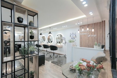 A modern and stylish beauty salon interior featuring wooden accents and a sleek design. The salon has a spacious layout with multiple salon chairs and mirrors under warm pendant lighting. Plants are used as decorative elements, adding a fresh and inviting atmosphere. The reception area has a wooden backdrop with the salon's name displayed, and there's a mix of contemporary furniture and decor.