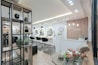 A modern and stylish beauty salon interior featuring wooden accents and a sleek design. The salon has a spacious layout with multiple salon chairs and mirrors under warm pendant lighting. Plants are used as decorative elements, adding a fresh and inviting atmosphere. The reception area has a wooden backdrop with the salon's name displayed, and there's a mix of contemporary furniture and decor.