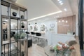 A modern and stylish beauty salon interior featuring wooden accents and a sleek design. The salon has a spacious layout with multiple salon chairs and mirrors under warm pendant lighting. Plants are used as decorative elements, adding a fresh and inviting atmosphere. The reception area has a wooden backdrop with the salon's name displayed, and there's a mix of contemporary furniture and decor.