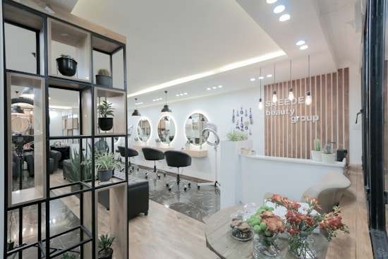 A modern and stylish beauty salon interior featuring wooden accents and a sleek design. The salon has a spacious layout with multiple salon chairs and mirrors under warm pendant lighting. Plants are used as decorative elements, adding a fresh and inviting atmosphere. The reception area has a wooden backdrop with the salon's name displayed, and there's a mix of contemporary furniture and decor.
