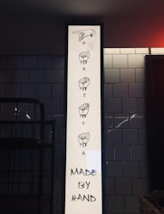 A vertical white sign displays illustrations of sign language hand gestures spelling out the word 'PASTA'. Below the illustrations, black text reads 'MADE BY HAND'. The sign is set against a background of white subway tiles, and there is a darkened area that appears to be indoor lighting ambience.