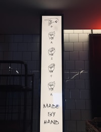 A vertical white sign displays illustrations of sign language hand gestures spelling out the word 'PASTA'. Below the illustrations, black text reads 'MADE BY HAND'. The sign is set against a background of white subway tiles, and there is a darkened area that appears to be indoor lighting ambience.