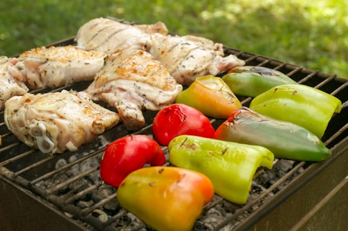 Several pieces of seasoned chicken alongside colorful bell peppers are grilling on a barbecue under sunny conditions. The chicken is lightly charred, and the bell peppers display vibrant shades of red, orange, and green. Charcoal can be seen beneath the grill, and the scene seems to be outdoors, indicated by the blurred green background suggesting grass or garden foliage.