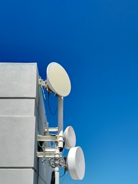 A set of satellite dishes mounted on the side of a building against a clear blue sky. The dishes are white and round, attached to poles and connected with cables. The building wall is light gray with distinct lines dividing the sections.