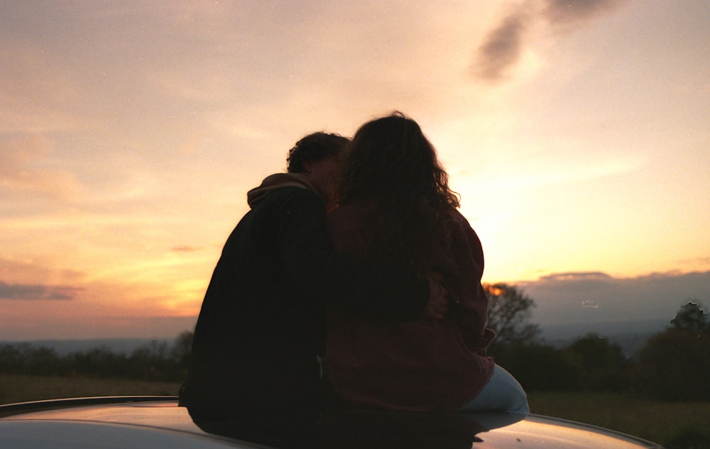 two people sitting on the hood of a car at sunset
