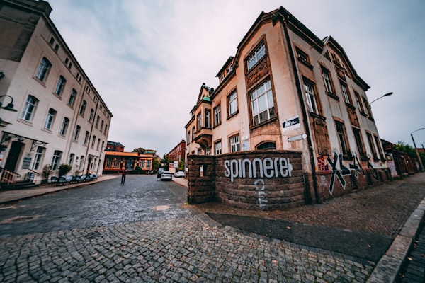 An urban street scene captured with a wide-angle perspective, featuring old architectural buildings with graffiti on their walls. The street is paved with cobblestones, leading to several parked cars and a person walking in the distance. Overcast skies and muted colors add to the image.