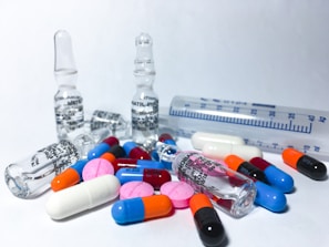 Various colorful pills and ampoules are arranged on a white surface, alongside a transparent measuring tube. The pills include capsules and tablets in colors such as blue, orange, pink, red, and black. The ampoules are made of clear glass, indicating a medical or pharmaceutical theme.