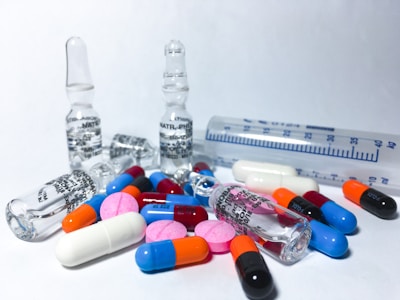 Various colorful pills and ampoules are arranged on a white surface, alongside a transparent measuring tube. The pills include capsules and tablets in colors such as blue, orange, pink, red, and black. The ampoules are made of clear glass, indicating a medical or pharmaceutical theme.
