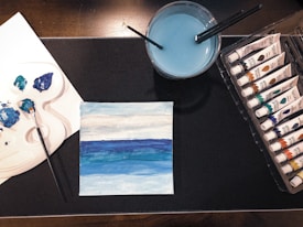 A painting setup with a small canvas depicting a blue gradient landscape placed on a dark surface. Several tubes of acrylic paint are neatly organized in a plastic tray to the right. A container of blue water with brushes is positioned towards the top center, while a palette with a mix of blue paint and a brush lies on the left.